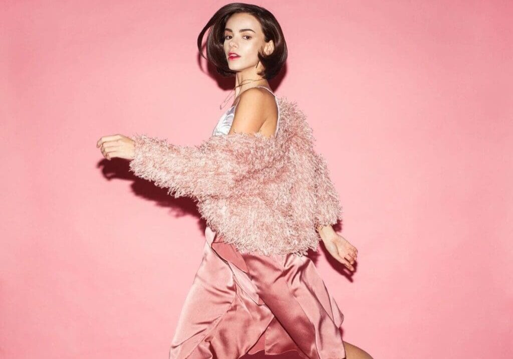 A woman wearing a fluffy pink jacket and satin pink skirt walks in front of a solid pink background.