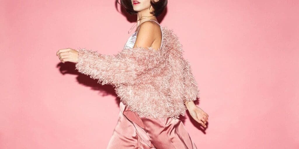 A woman wearing a fluffy pink jacket and satin pink skirt walks in front of a solid pink background.