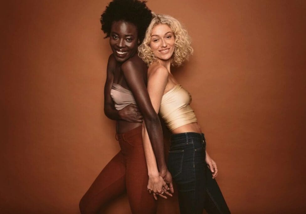 Two women stand side by side against a brown background, smiling and holding hands. One has dark skin and curly hair; the other has light skin and blonde curly hair.