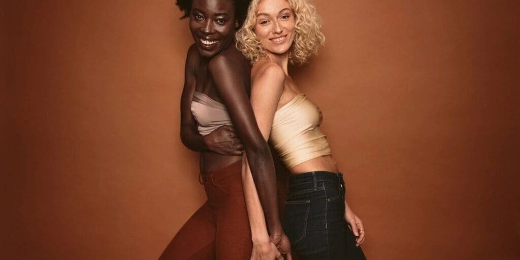 Two women stand side by side against a brown background, smiling and holding hands. One has dark skin and curly hair; the other has light skin and blonde curly hair.