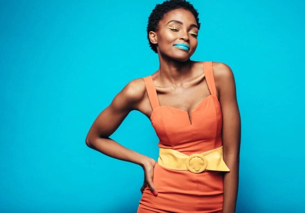 Woman in an orange dress with a wide yellow belt stands against a blue background, wearing blue lipstick and yellow eyeshadow, with one hand on her hip and eyes closed.