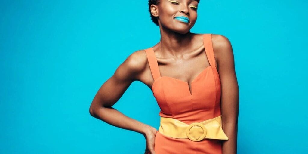 Woman in an orange dress with a wide yellow belt stands against a blue background, wearing blue lipstick and yellow eyeshadow, with one hand on her hip and eyes closed.
