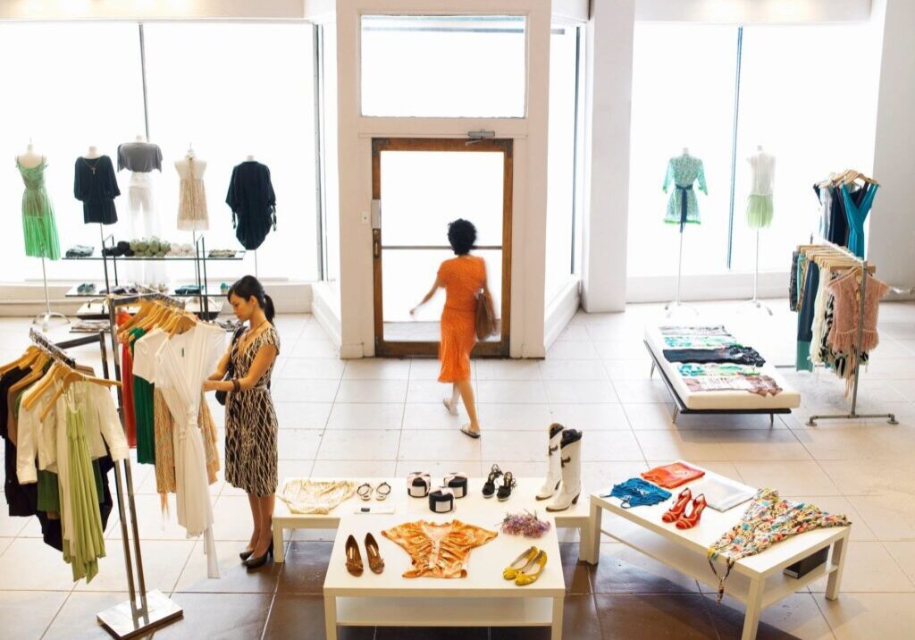 A woman browses clothing racks in a bright boutique while another woman in an orange dress walks out the front door.