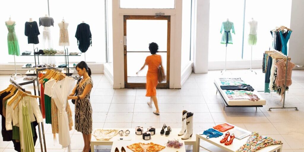 A woman browses clothing racks in a bright boutique while another woman in an orange dress walks out the front door.