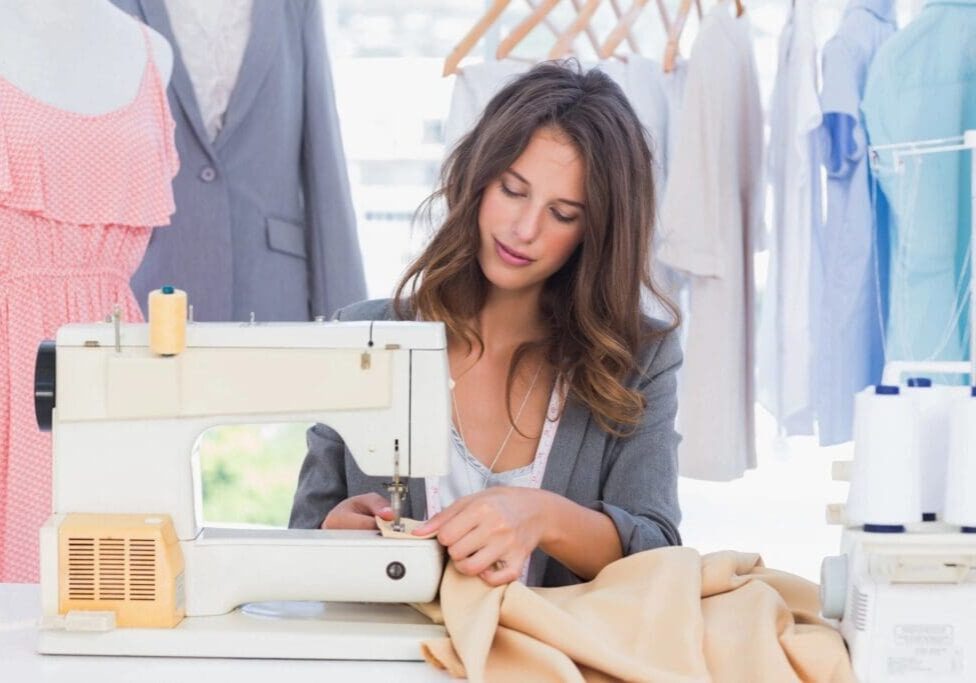 A woman sits at a sewing machine, stitching beige fabric, with clothing hanging in the background.