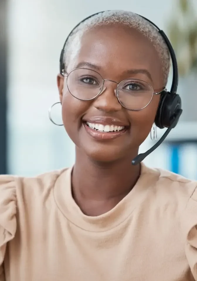 A person wearing glasses, a headset, and a light-colored top smiles while seated in an office environment.